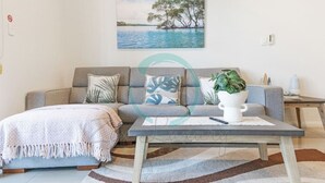 En stue med en grå sofa, et sofabord med en plante, og et maleri af en sø omgivet af træer.