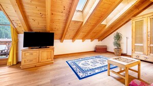 Una habitación con techo de madera inclinado, un televisor sobre un mueble de madera, un tapete azul estampado y una mesa de madera.