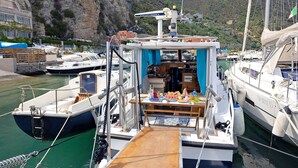 A boat with a small deck area, a table with food, and a view of other boats and a rocky cliff in the background. - Aloha Jak's Boat with Sea View, Wi-Fi, and Air Conditioning (Alassio)