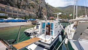 A marina with boats docked, a building with multiple windows, and a rocky hillside in the background. - Aloha Jak's Boat with Sea View, Wi-Fi, and Air Conditioning (Alassio)