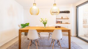 Un coin repas avec une table en bois, des chaises blanches, deux lampes suspendues, un vase avec des fleurs, une plante et une vue à travers une porte vitrée.