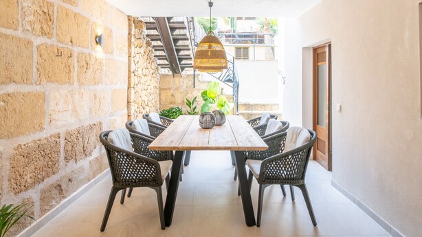 Un coin repas avec une table en bois et des chaises en osier, un mur en pierre et un escalier menant à l’extérieur.