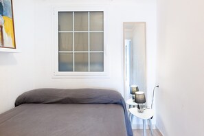 A bedroom with a bed, a window, a mirror, and a small table with a lamp.