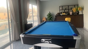 Une table de billard dans une pièce dotée de grandes fenêtres, d’un ventilateur de plafond et d’un comptoir d’accueil agrémenté d’objets décoratifs.