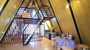 Un intérieur spacieux, doté d’un toit en pente et d’un escalier en bois menant à un niveau supérieur. On aperçoit un coin repas avec une table et des tabourets.