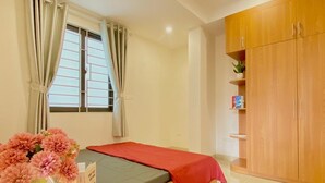 A bedroom with a bed, curtains, a wooden wardrobe, and a small table with a vase of flowers. - Quan Nhan Apartment (Hanoi)