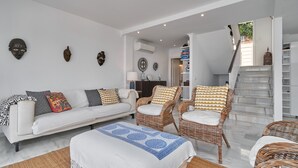Een woonkamer met een witte bank, rieten stoelen, een bed met een blauw beddegoed met patroon, en een trap naar een tweede verdieping.