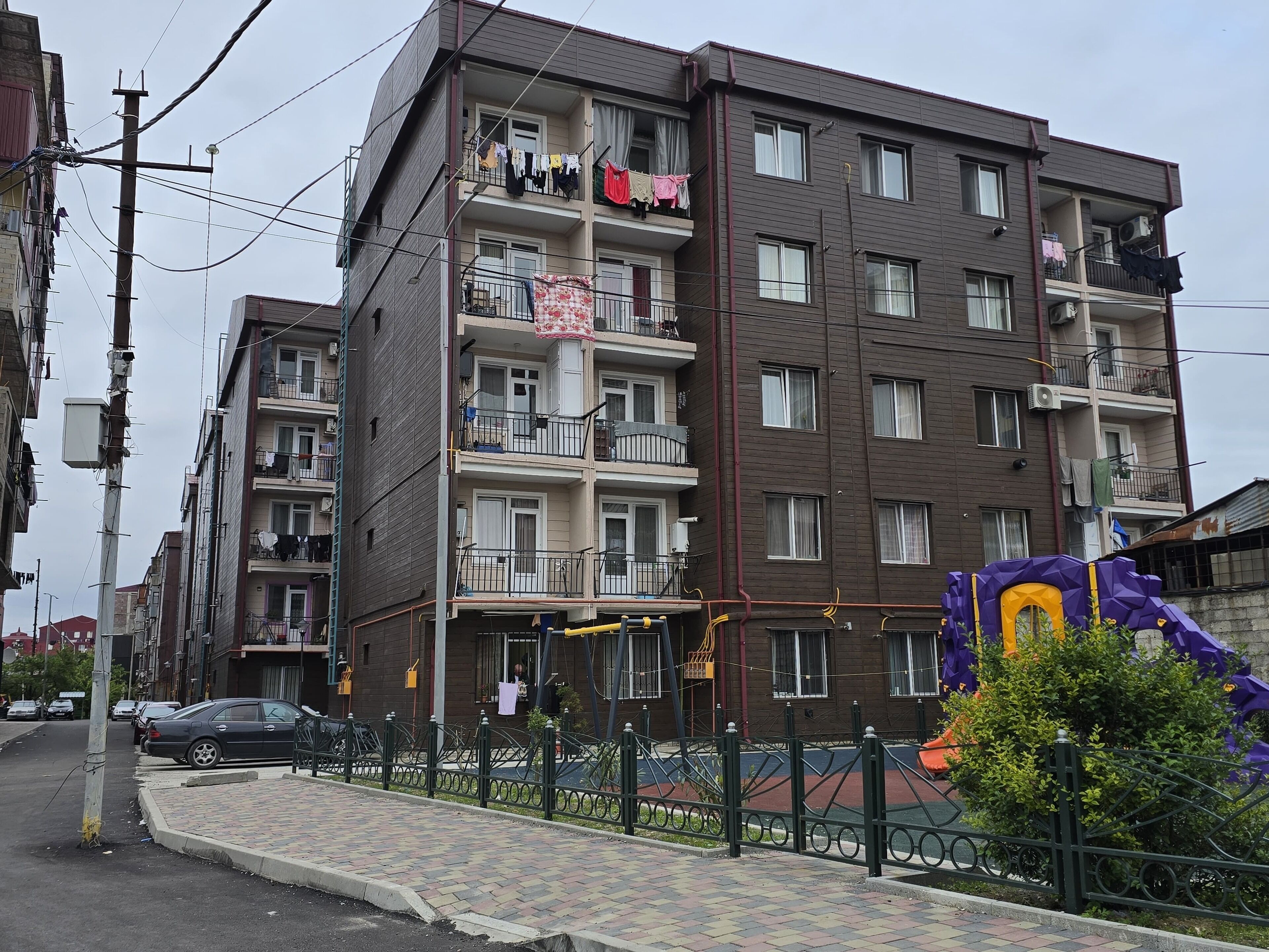 A multi-story residential building with balconies, a playground, and a paved sidewalk.