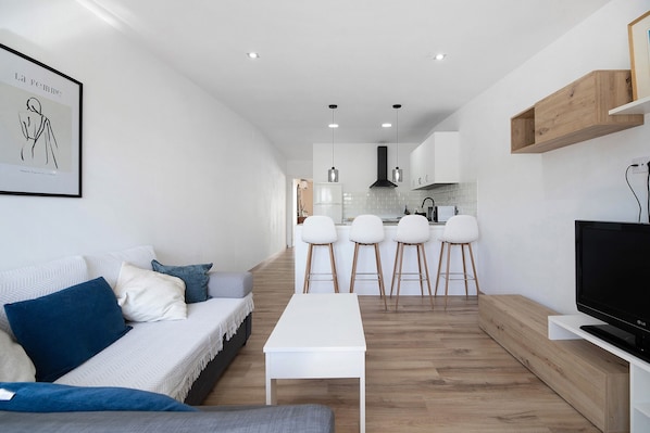Un salon moderne avec un canapé, une table basse, un téléviseur et une cuisine en arrière-plan.