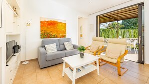 A modern living room with a grey sofa, a white coffee table, and a beige rocking chair. There is a painting on the wall, a TV on a stand, and a large window with a view of greenery. - Villa 'Alfonsa' with Mountain View, Wi-Fi and Air Conditioning (pollensa)