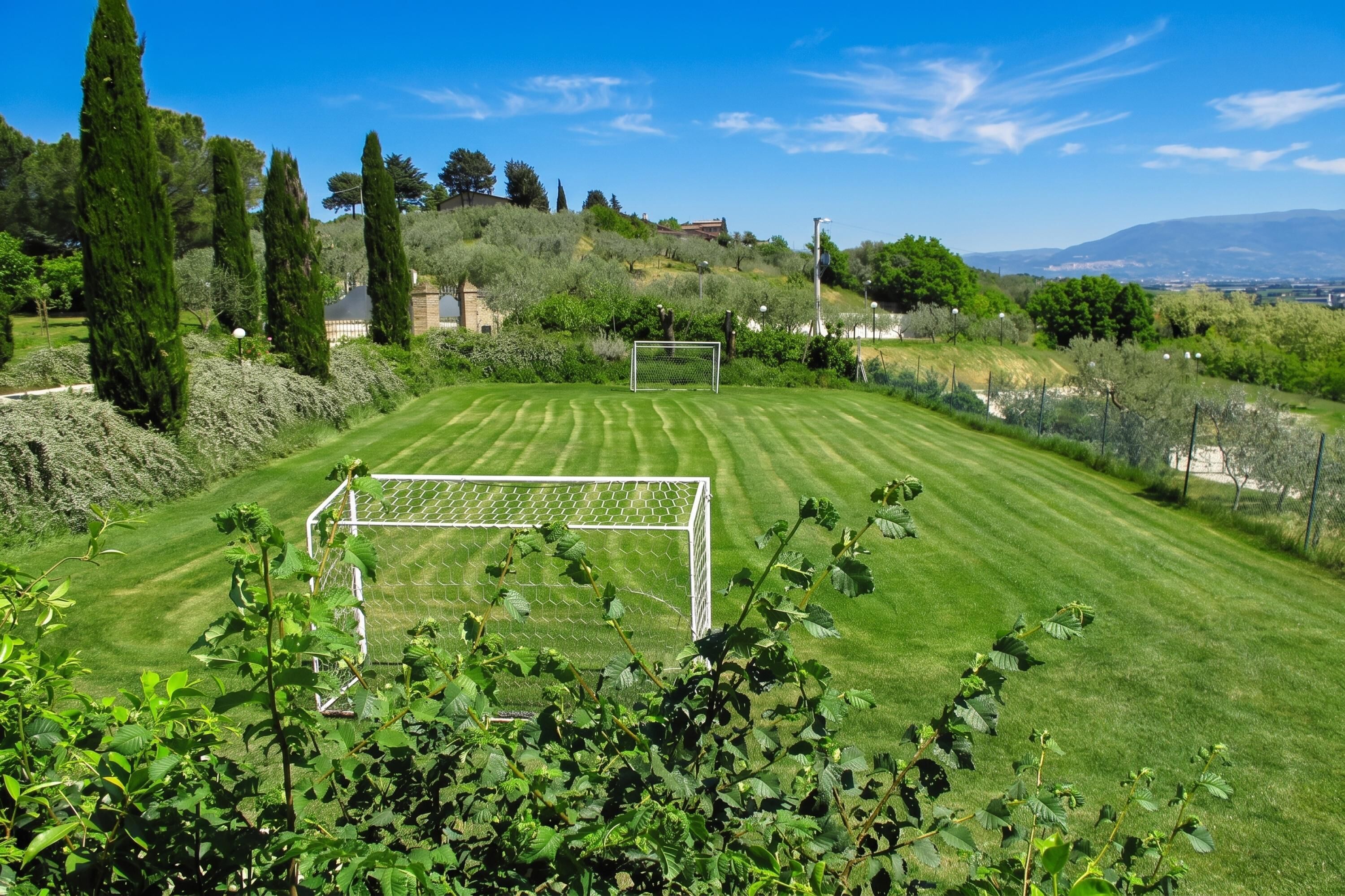 A well-maintained grassy area with a soccer goal, surrounded by greenery and hills in the background.
