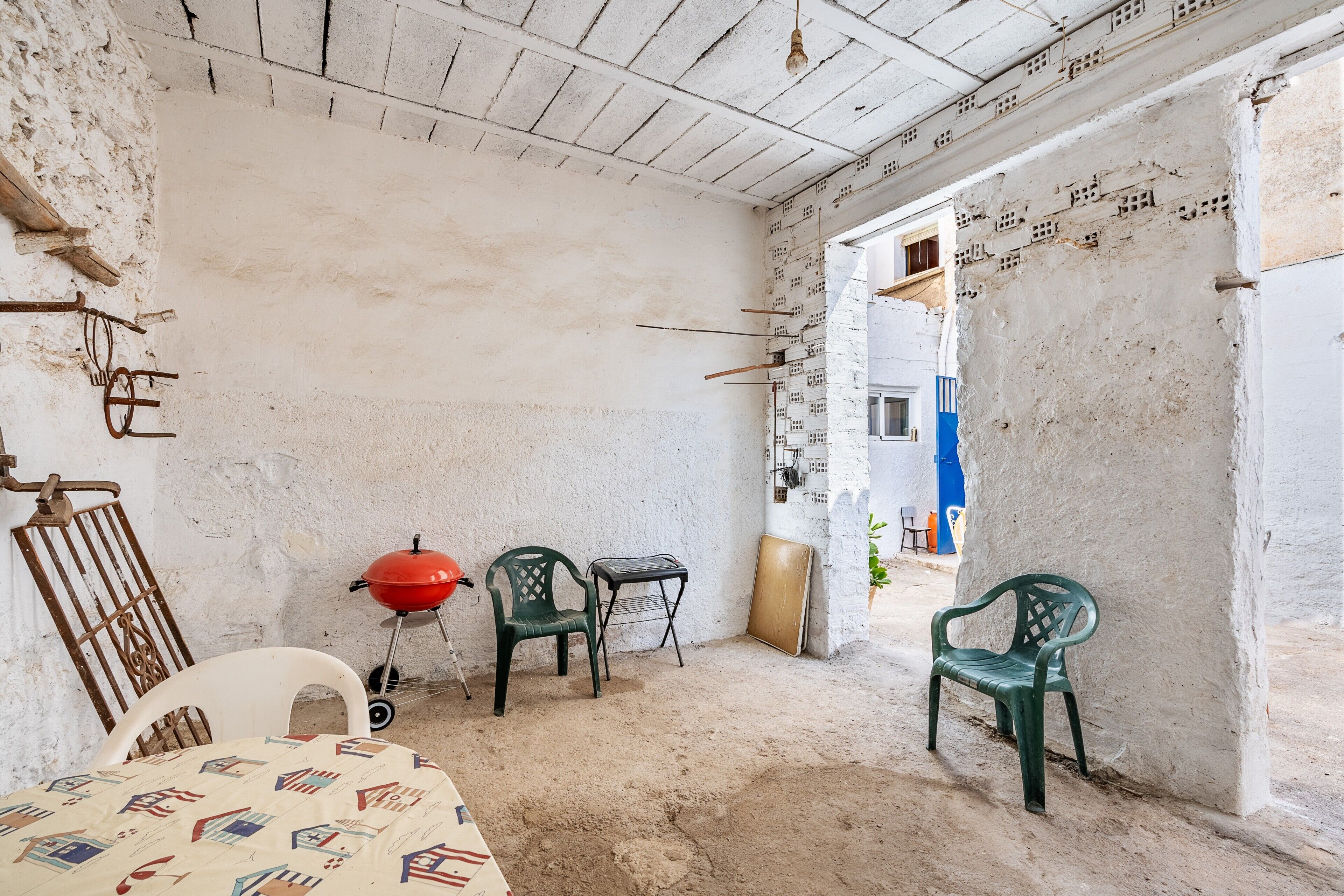 Une pièce avec un plafond blanc, une table recouverte d’une nappe à motifs, deux chaises en plastique vertes, un barbecue rouge et, au fond, une porte bleue.