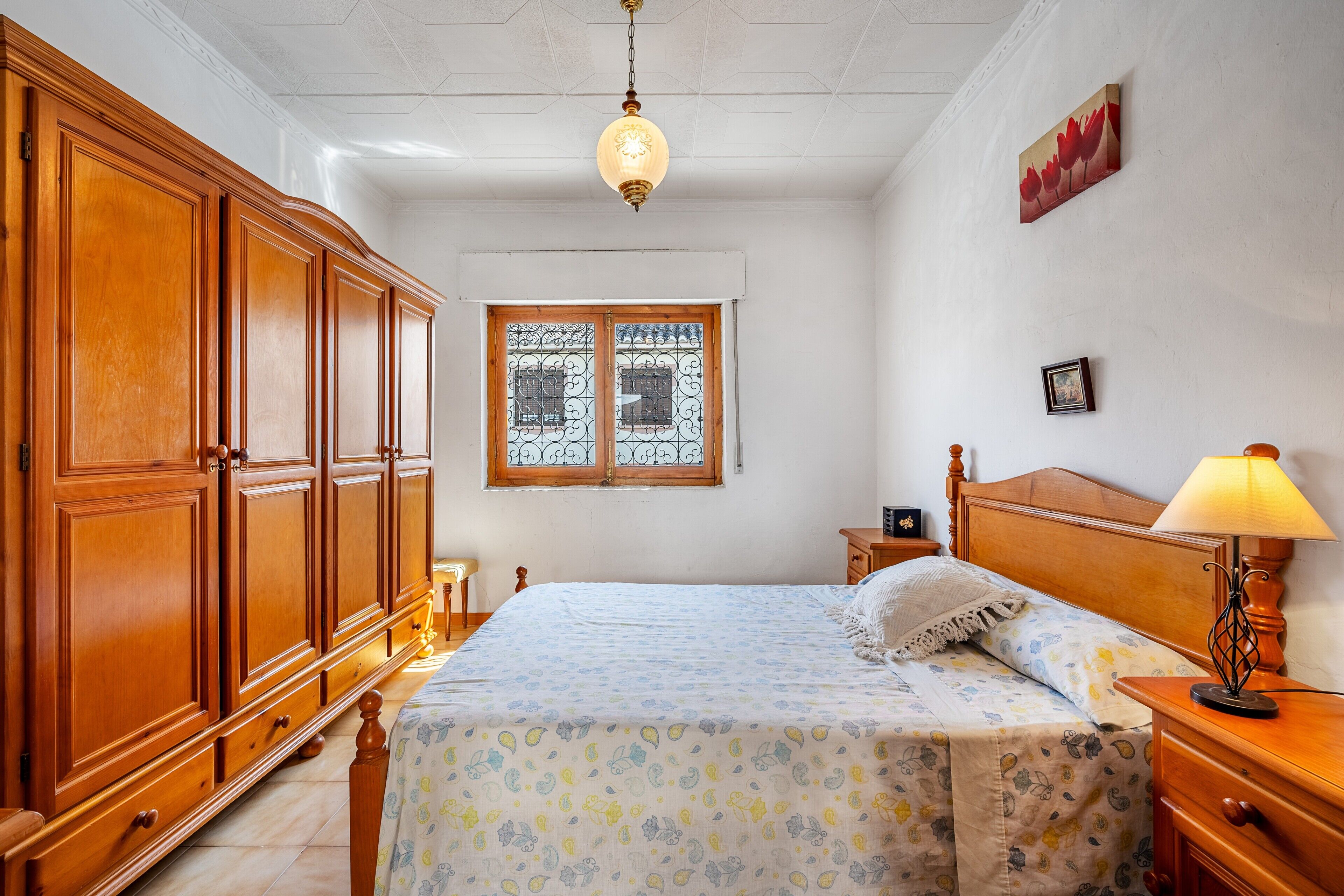 A bedroom with a large wooden wardrobe, a bed with a floral bedspread, a nightstand with a lamp, and a window with decorative iron bars.