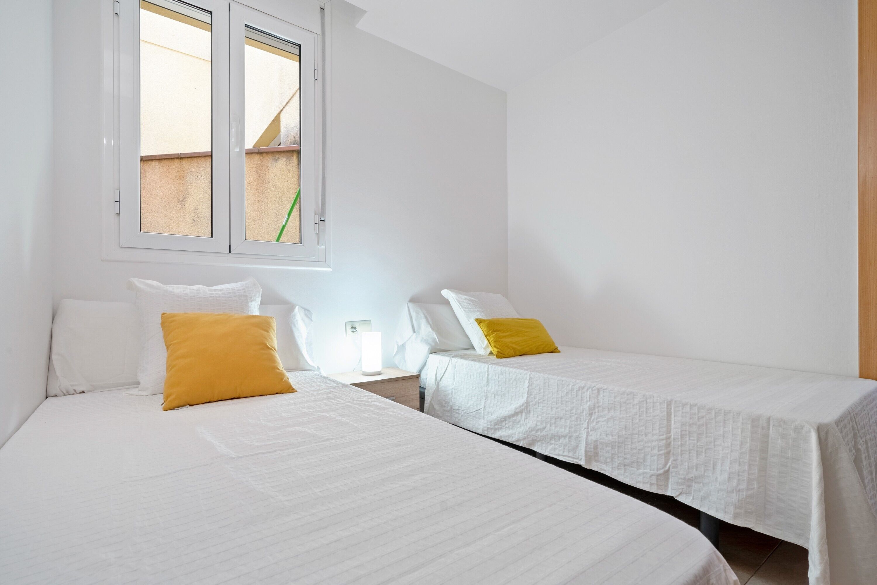 Una habitación con dos camas, ropa de cama blanca, una ventana con vistas y una mesita de noche con una lámpara.
