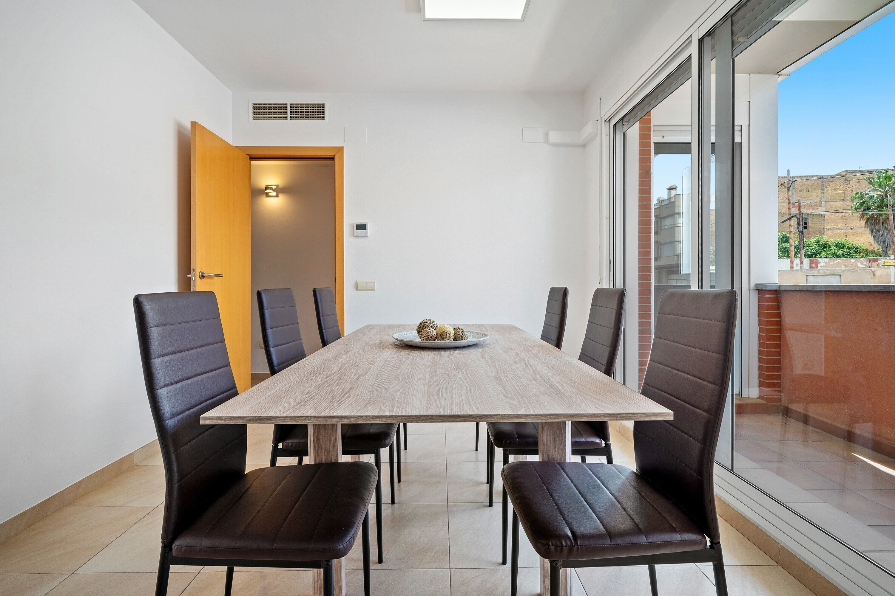 Un espace repas avec une table en bois et des chaises noires, une porte donnant sur une autre pièce et une vue à travers de grandes fenêtres.