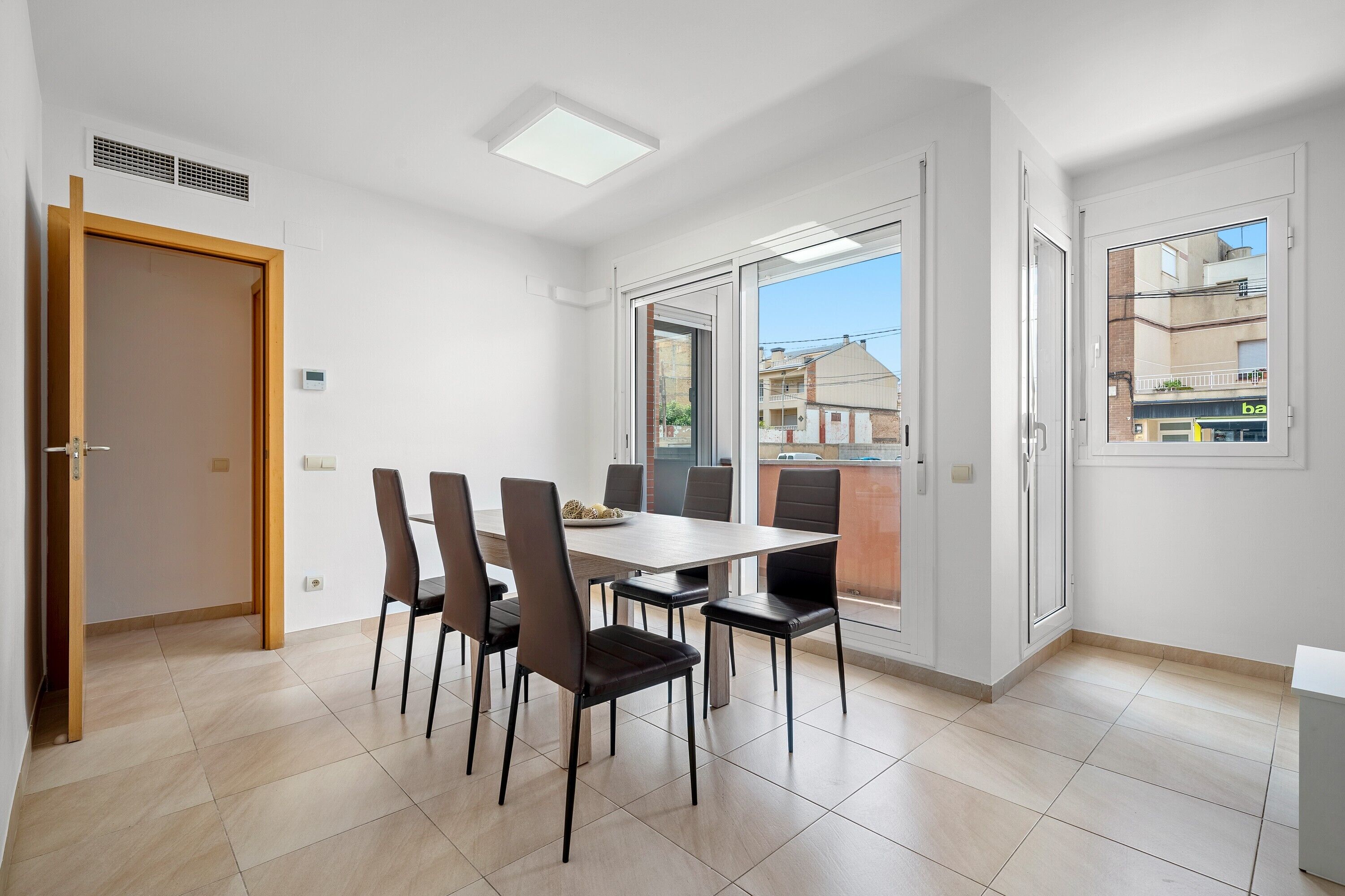 Un comedor moderno con una mesa y sillas, un vidrio corredero hacia un balcón y vistas de edificios.