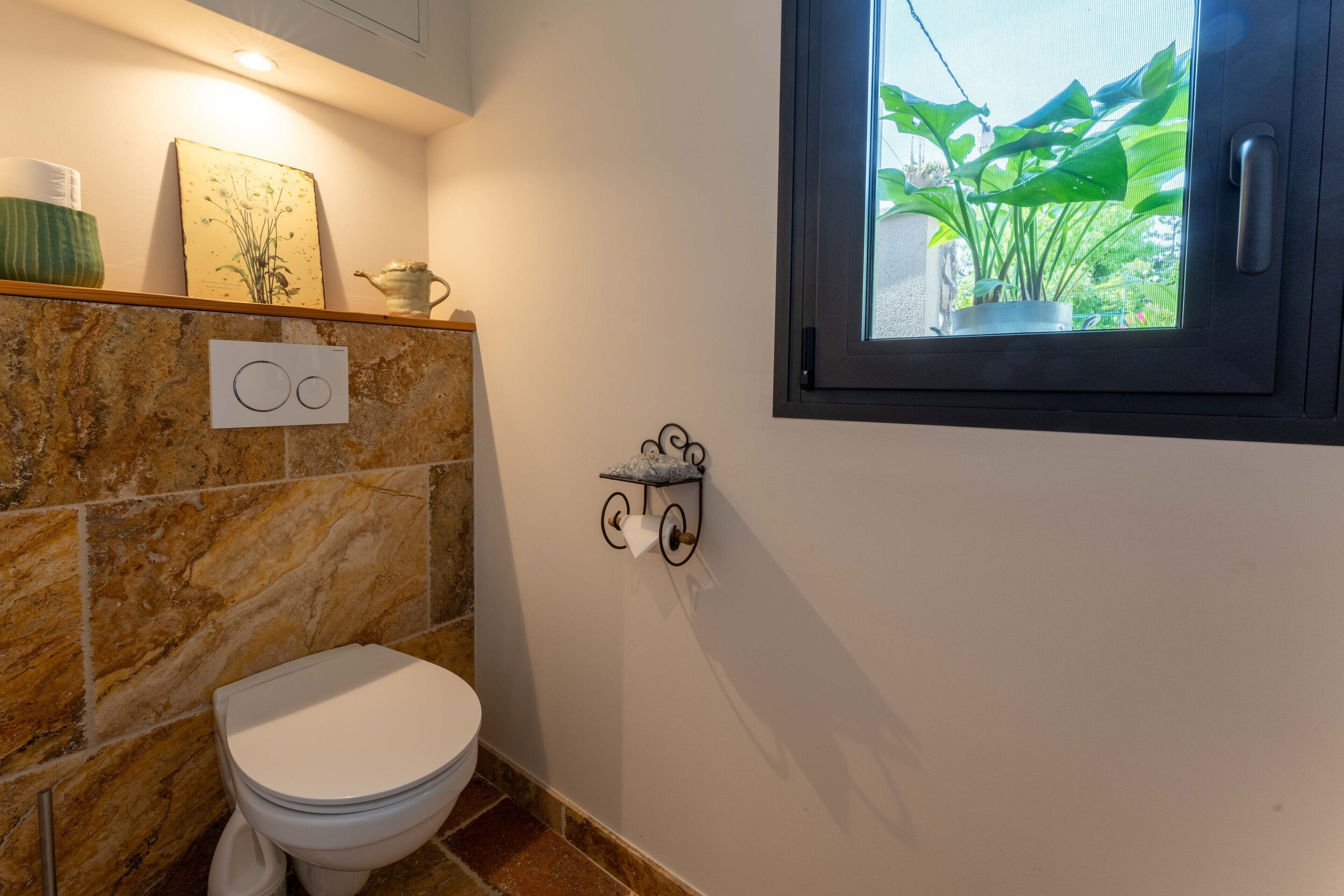 A bathroom with a toilet, a wall-mounted toilet paper holder, a window with a view of greenery, and a decorative item on the wall.