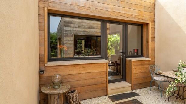 Un mur extérieur en bois, une grande porte-fenêtre coulissante, une petite table ronde avec un vase en verre et une chaise en métal.