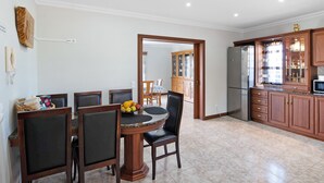 A dining area with a round table, chairs, and a fruit bowl. There is a kitchen with wooden cabinets and a refrigerator.
