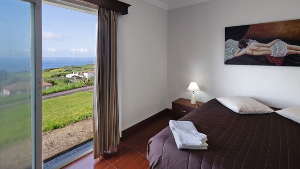 A hotel room with a bed, a painting, a nightstand, a lamp, and a view of the ocean through a window.