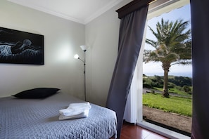 A bedroom with a bed, a painting, a lamp, and a view of palm trees through a window. - Villa 'Vivenda Do Monte' with Sea View, Shared Garden and Wi-Fi (Achadinha)