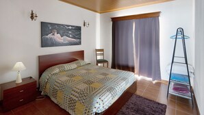 A bedroom with a bed, a chair, a painting, a lamp, a window with curtains, and a drying rack. - Holiday Home 'Casa Do Monte' with Sea View, Shared Garden and Wi-Fi (Achadinha)