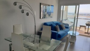 Un salon moderne avec une table à manger en verre, des chaises blanches, un canapé bleu et une grande fenêtre donnant sur la plage.
