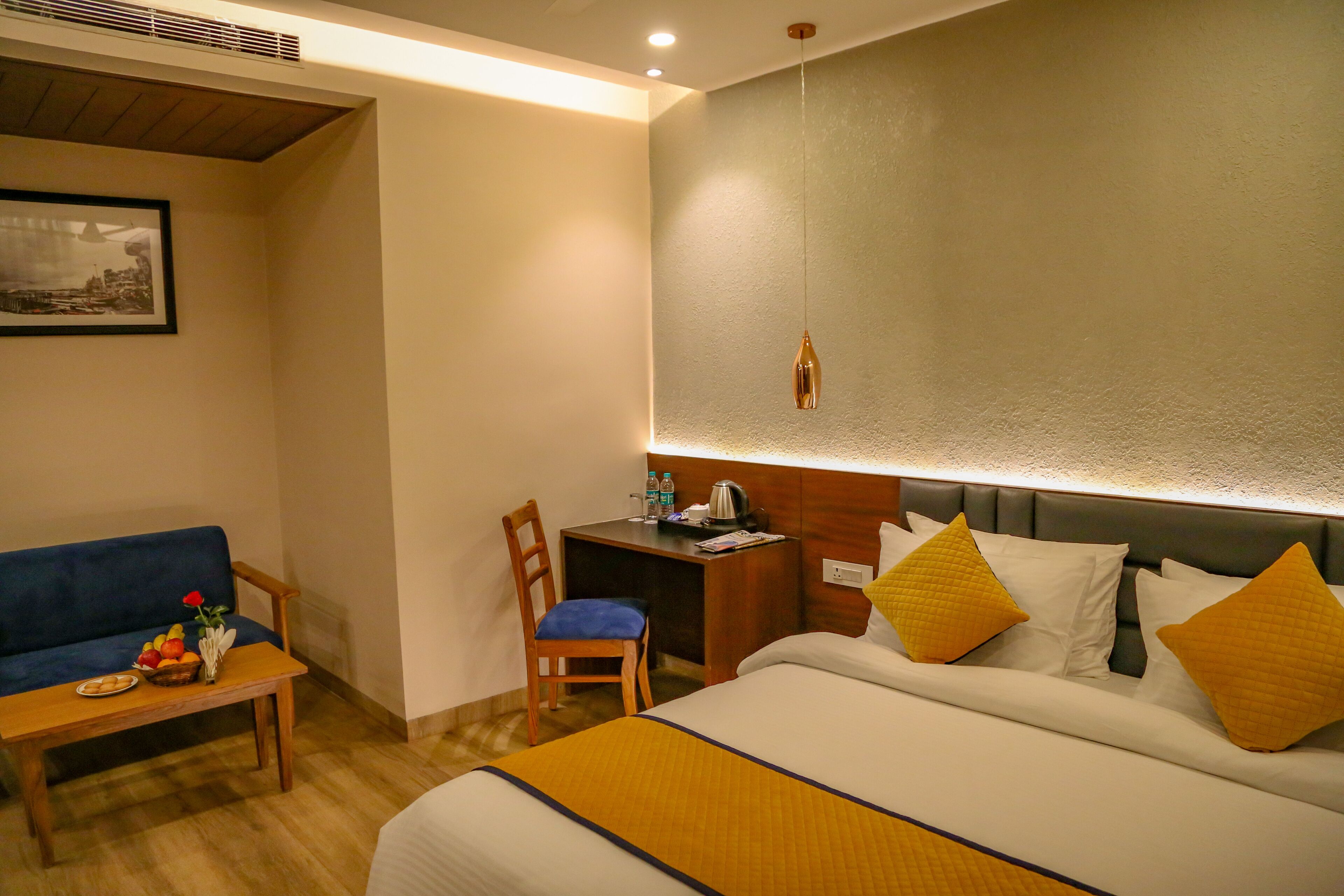 A hotel room with a bed, a desk, a chair, a sofa, and a small table with a fruit bowl and a vase.