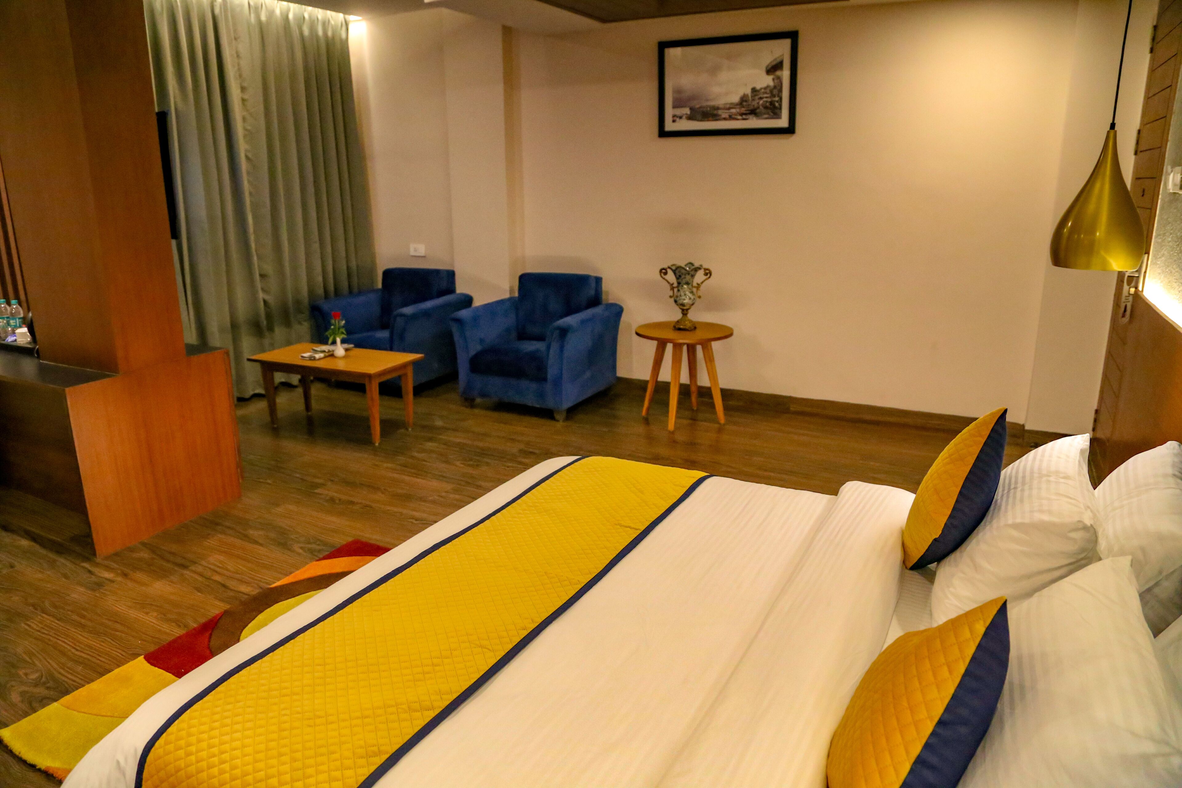 A hotel room with a large bed, two blue armchairs, a small round table, and a wooden desk.