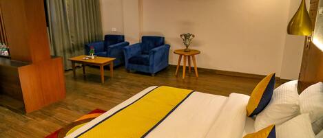 A hotel room with a large bed, two blue armchairs, a small round table, and a wooden desk.