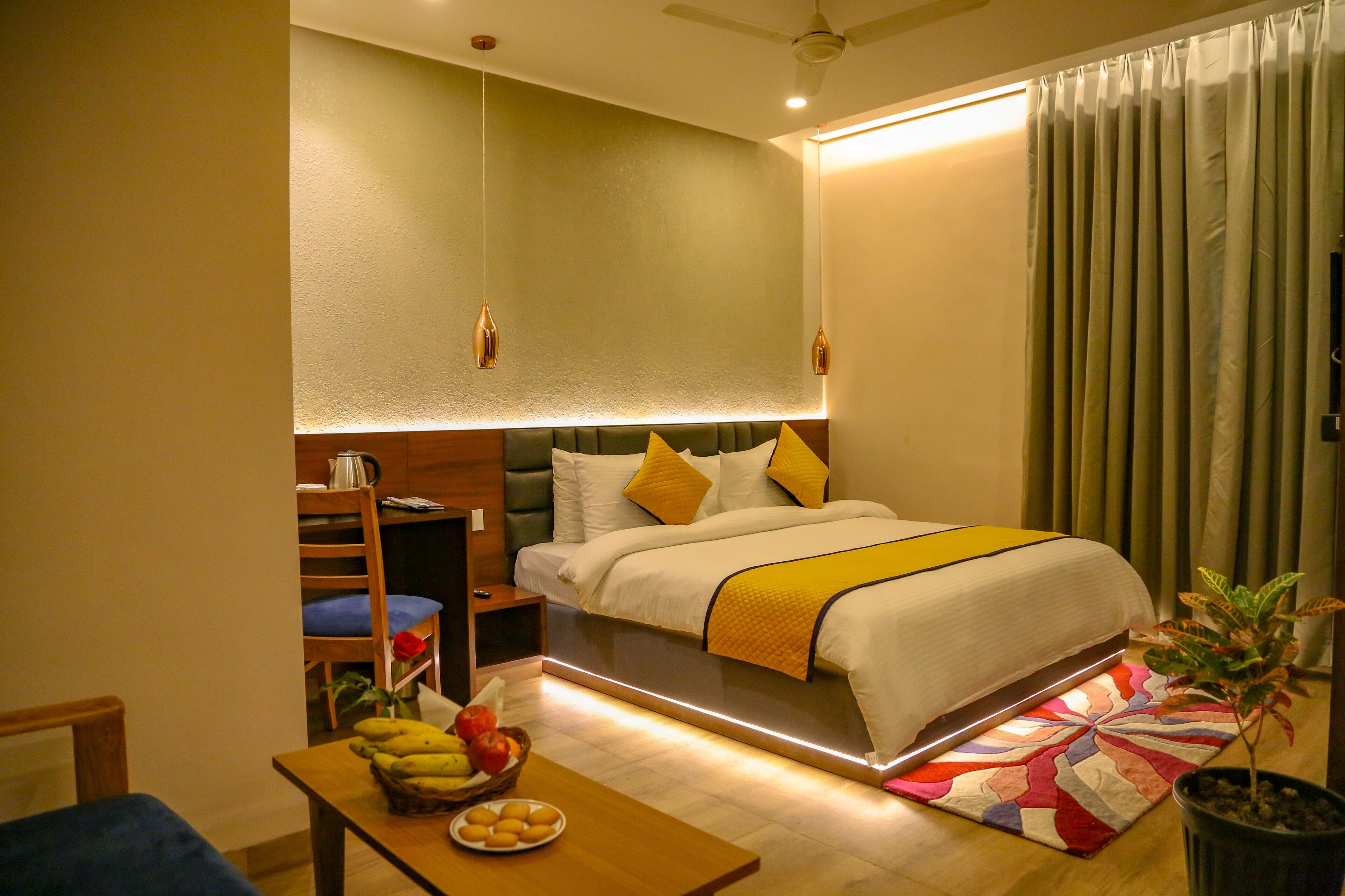 A hotel room with a bed, a desk with a fruit bowl, a chair, and a potted plant.