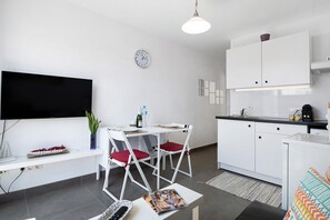 A modern kitchen with white cabinets, a dining area with white chairs, a flat-screen TV, and a clock on the wall.