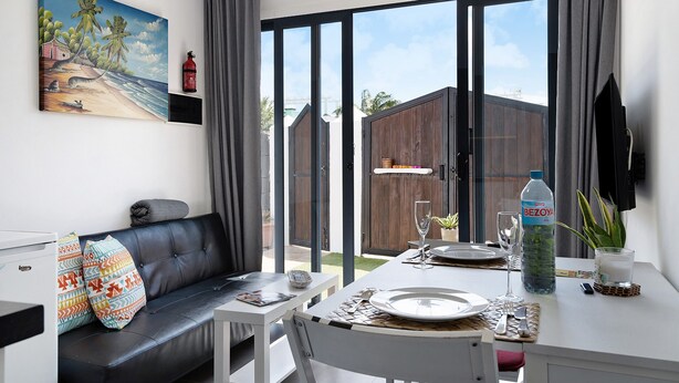 A dining area with a table set for two, a sofa with patterned cushions, a painting on the wall, and a view of the outside.