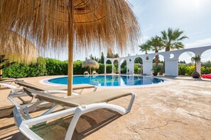 Un área de piscina con sillas de descanso, una sombrilla de paja y una estructura arqueada blanca.