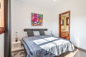 Un dormitorio con cama, mesita de noche, lámpara, un cuadro en la pared y una puerta que da a otra habitación.