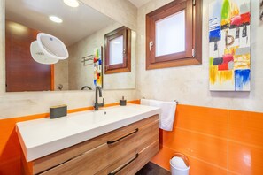 Un baño con lavamanos blanco, tocador de madera, azulejos naranjas, un espejo grande y una ventana con marco de madera.