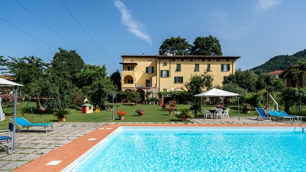 A pool area with a rectangular pool, lounge chairs, and a building with a yellow facade.