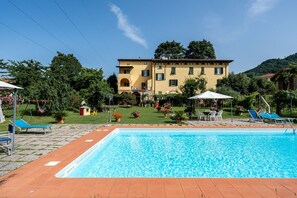A pool area with a rectangular pool, lounge chairs, and a building with a yellow facade.