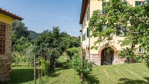 A building with a stone base and yellow upper walls, surrounded by a grassy area with trees and a vineyard.