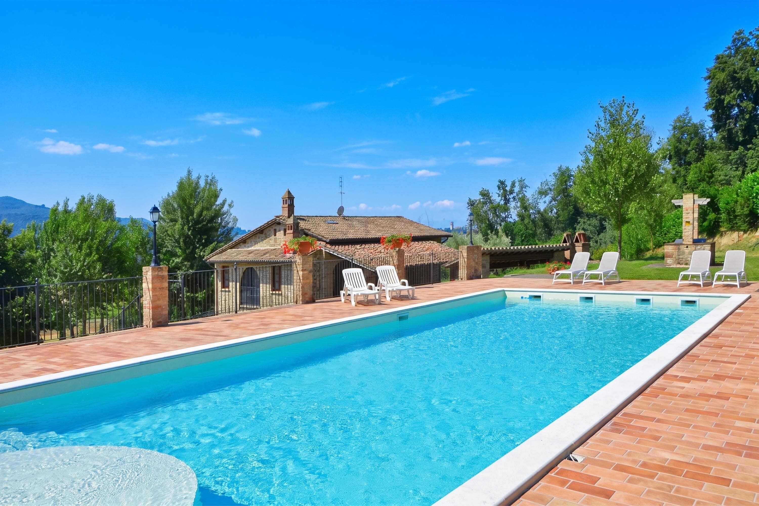 A rectangular swimming pool with a tiled deck, surrounded by white lounge chairs, a small building with a tiled roof, and a stone wall.