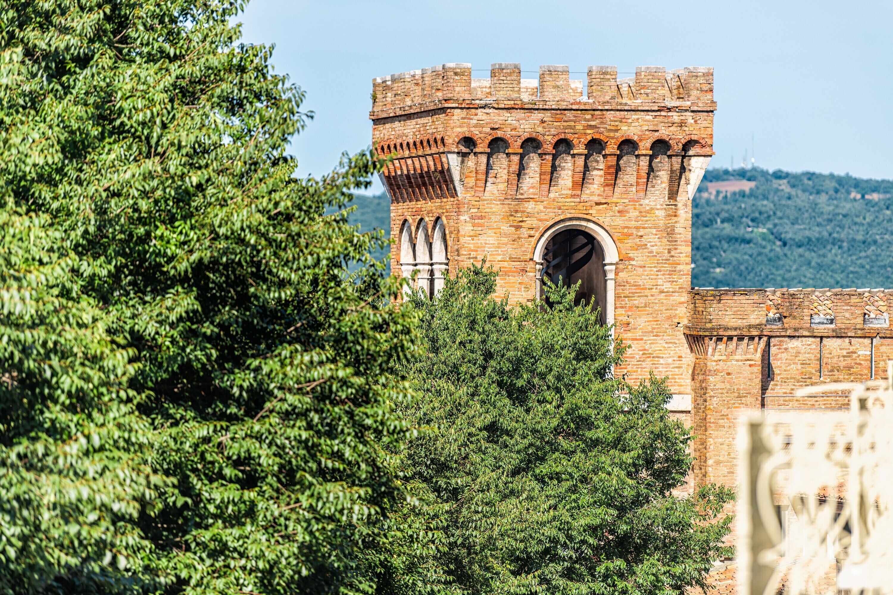 A historic brick tower with arched windows and a crenellated top, surrounded by dense green foliage.