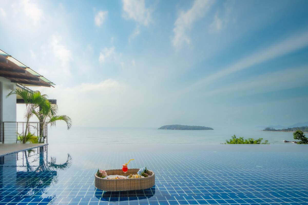 An infinity pool with a wicker basket containing a drink and snacks, overlooking a calm sea and distant island.