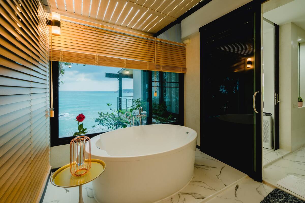 A modern bathroom with a large bathtub, a small table with a vase, and a view of the sea.