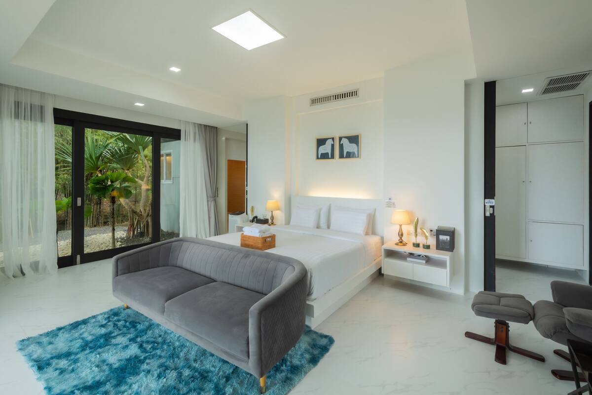 A modern hotel room with a large bed, a grey sofa, a blue rug, and a view of palm trees through the glass door.