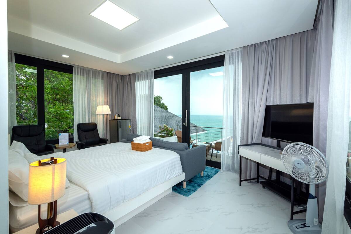 A modern hotel room with a large bed, a desk with a flat-screen TV, a desk fan, and a balcony with a view of the sea.
