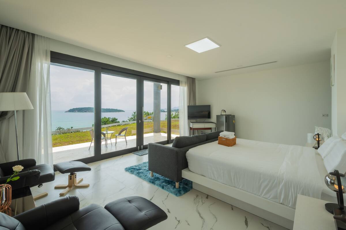 A modern hotel room with a large bed, a TV, and a balcony with a view of the sea.