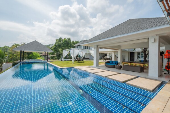 Una piscina con solárium empedrado, un porche cubierto y vista a una casa al fondo.