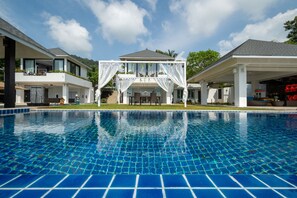 Une piscine entourée d’une terrasse carrelée, bordée de maisons modernes dotées de balcons et agrémentée d’un espace couvert avec un auvent blanc.
