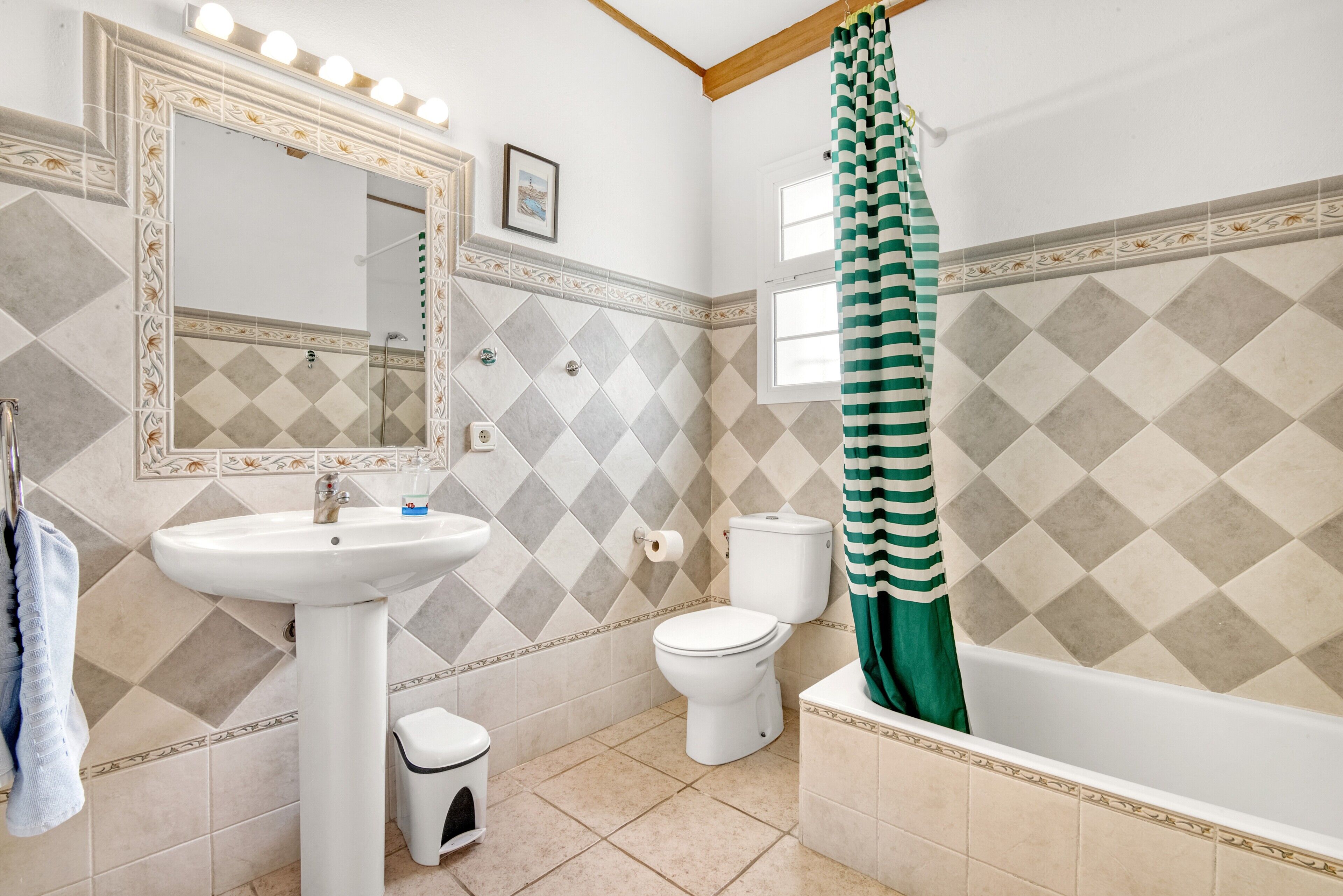 Un baño con lavabo, inodoro y bañera, con azulejos estampados y un espejo con iluminación.