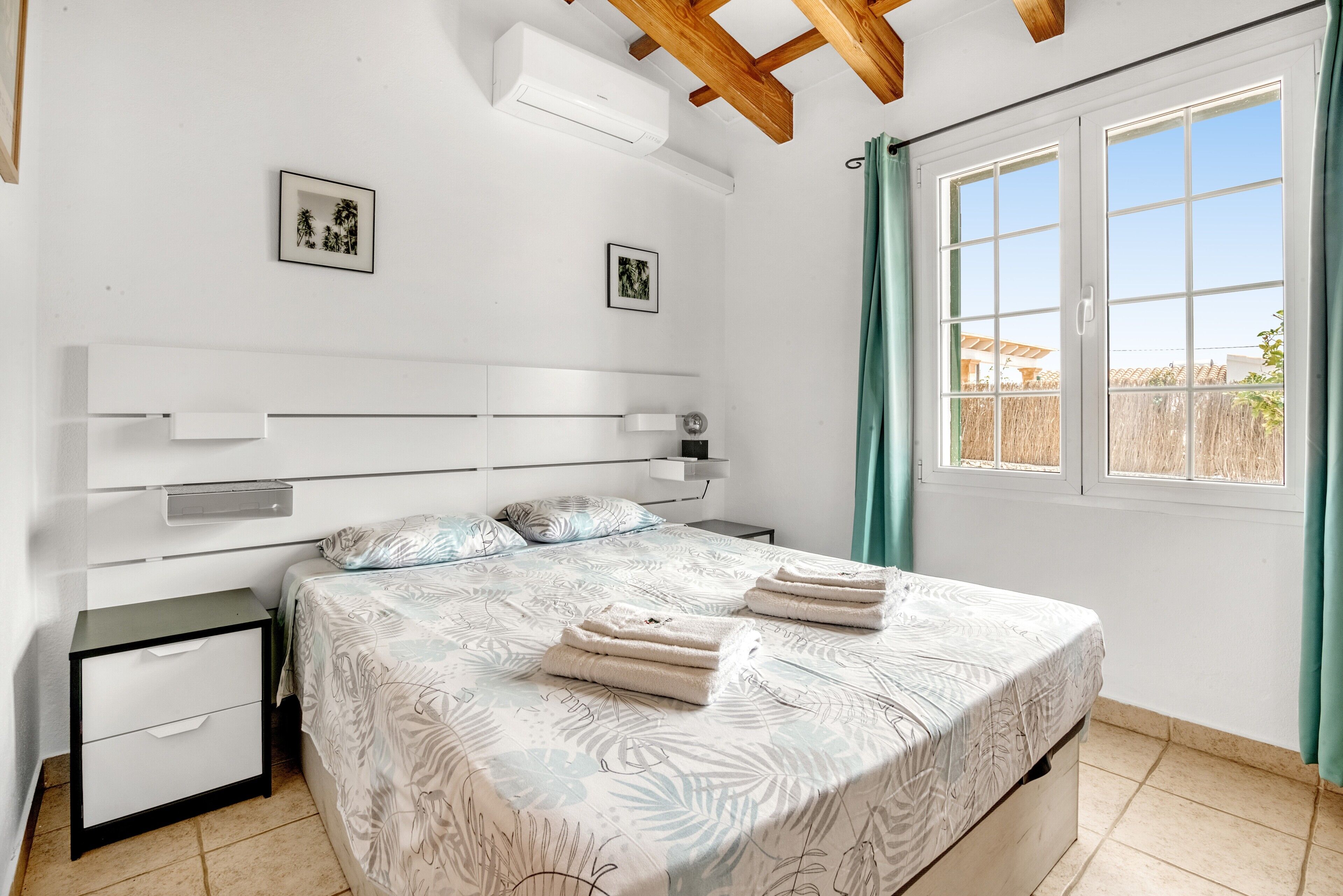 Ein Schlafzimmer mit Bett, Nachttischen, einem Fenster mit Vorhängen und einer Wand-Klimaanlage.